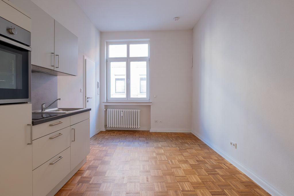Leere, helle Küche mit weißen Schränken, Backofen und Spüle links, Holzboden und Fenster mit Heizkörper an der Rückwand.