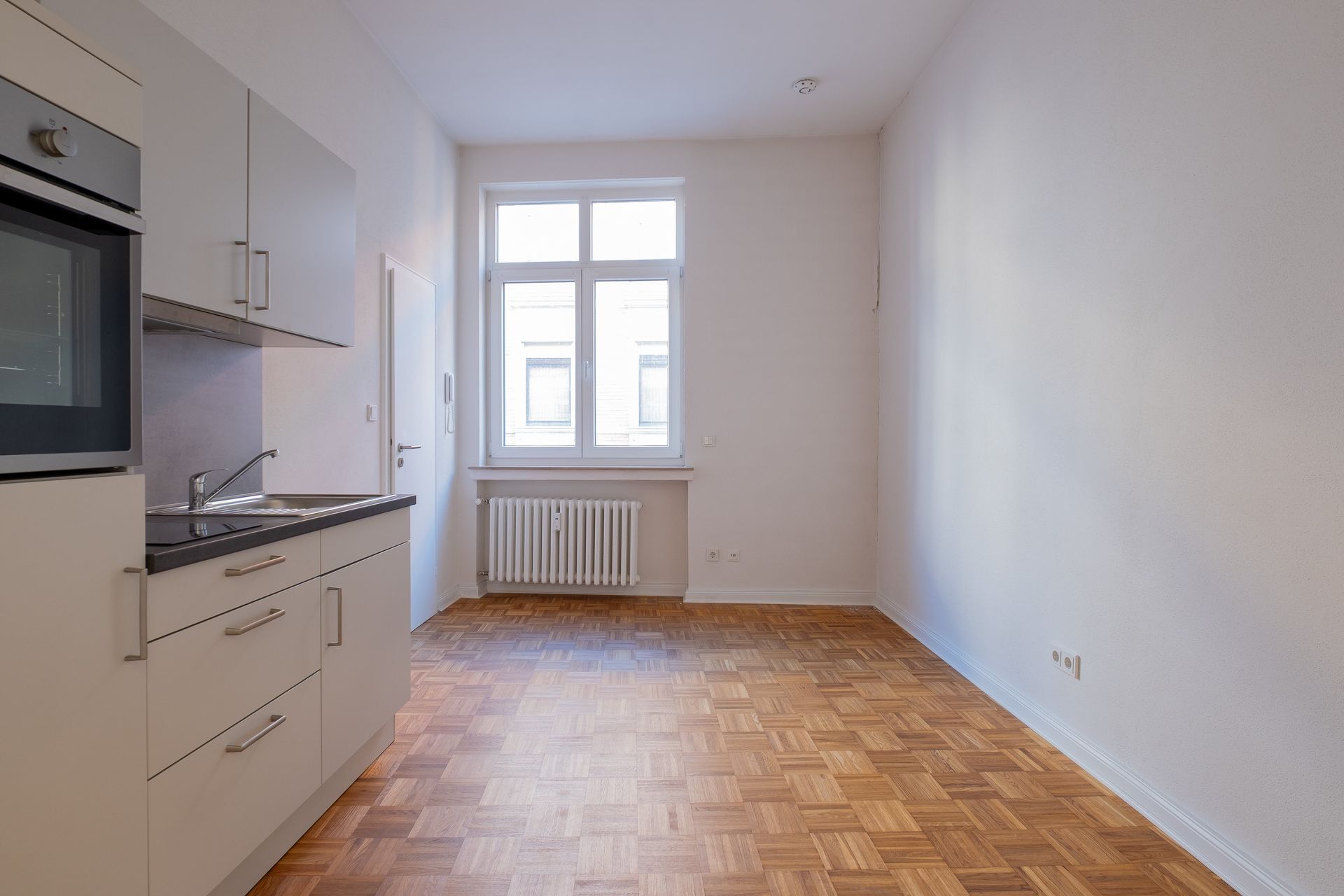 Leere, helle Küche mit weißen Schränken, Backofen und Spüle links, Holzboden und Fenster mit Heizkörper an der Rückwand.